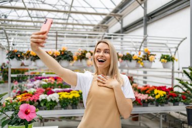 Neşeli bir Kafkasyalı kadın çiçekçi dükkanında selfie çekiyor, etrafında renkli çiçekler ve bitkiler var, bir masada..