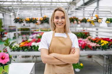 Gülümseyen bir çiçekçi dükkanında kollarını kavuşturarak gururla durur. Etrafı canlı çiçekler ve yeşilliklerle çevrilidir..