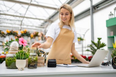 Çiçekçi dükkanındaki beyaz bir kadın dizüstü bilgisayarda çalışıyor ve masa üzerindeki bitkileri düzenliyor..
