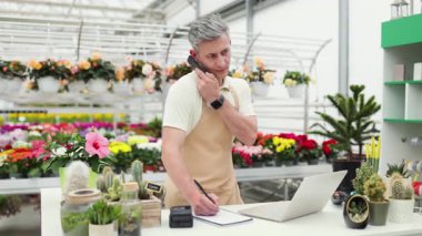 Önlüklü beyaz bir adam çiçekçinin tezgahında çalışıyor, dizüstü bilgisayar kullanıyor ve telefonda notlar alıyor..