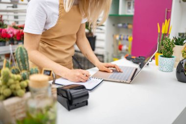 Diz üstü bilgisayarda çalışan ve çiçekçi tezgahında not defteri yazan beyaz bir kadın..