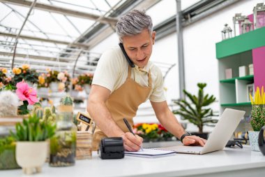 Beyaz bir erkek dükkan sahibi çiçekçi dükkanında bilgisayarında çalışırken telefonla sipariş veriyor..