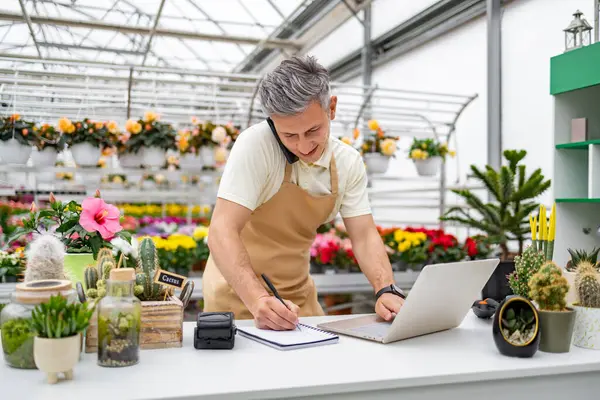 Çiçekçide çalışan beyaz bir adam telefonda not alıyor ve parlak bir günde dizüstü bilgisayar kullanıyor..