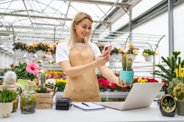 Gülümseyen bir çiçekçi dükkanında kaktüs resmi çeker ve satılık bitkilerin çeşitliliğini gösterir..