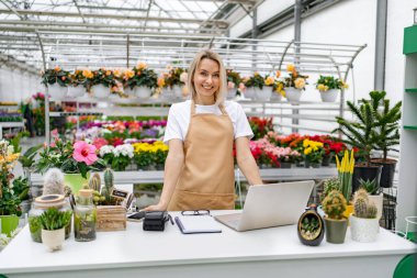 Beyaz bir kadın, renkli bitkilerle çevrili bir çiçekçide tezgahın arkasında duruyor. Kameraya bakıyor..