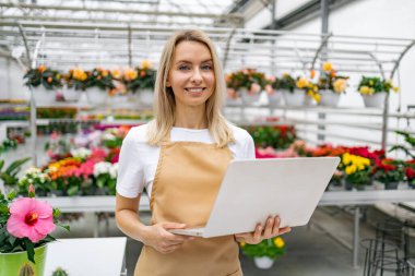Gülümseyen bir çiçekçi elinde dizüstü bilgisayarla dükkanında duruyor. Arkasında canlı çiçekler var..