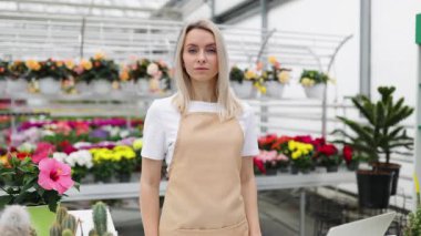 Gülümseyen bir çiçekçi dükkanında kollarını kavuşturarak gururla durur. Etrafı canlı çiçekler ve yeşilliklerle çevrilidir..