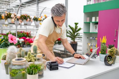 Önlüklü beyaz bir adam çiçekçinin tezgahında çalışıyor, dizüstü bilgisayar kullanıyor ve telefonda notlar alıyor..