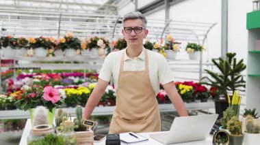 Çiçekçi dükkanında beyaz bir adam masanın arkasında çeşitli bitkilerle kameraya gülümsüyor..