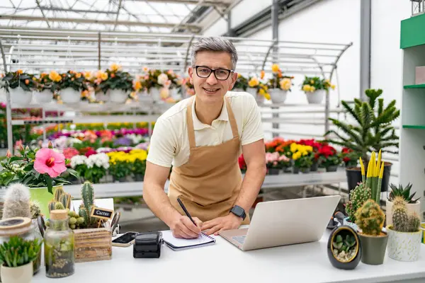 Kafkasyalı erkek bir çiçekçi, çeşitli bitki ve çiçeklerle çevrili bir tezgahtaki deftere yazıyor..