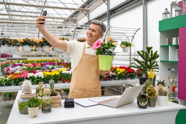 Çiçekçide beyaz bir adam saksı bitkisini tutarken selfie çekiyor..