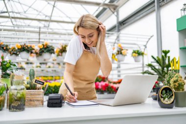 Çiçekçide beyaz bir kadın not defterine yazı yazıp dizüstü bilgisayar kullanarak telefonla sipariş veriyor..