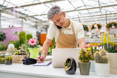 Çiçekçi dükkanında çalışan beyaz bir adam telefonla konuşuyor, deftere yazı yazıyor ve tezgahta dizüstü bilgisayar kullanıyor..