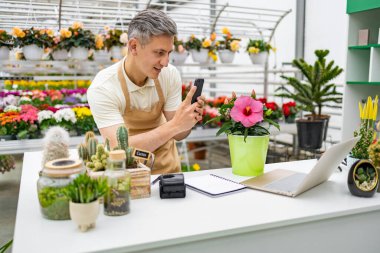 Beyaz bir adam dükkan ortamında bir çiçeğin fotoğrafını çekiyor. Masanın üzerinde çeşitli bitkiler ve dizüstü bilgisayarlar var..