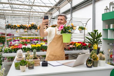 Kafkasyalı bir adam, bir çiçekçi, saksı çiçeği tutarken selfie çekiyor. Dükkan çeşitli bitkilerle dolu..