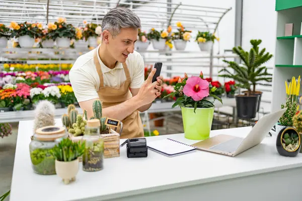Beyaz bir adam dükkan ortamında bir çiçeğin fotoğrafını çekiyor. Masanın üzerinde çeşitli bitkiler ve dizüstü bilgisayarlar var..