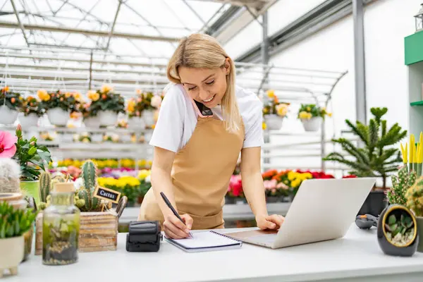 Beyaz bir kadın çiçekçiden sipariş alıyor, dizüstü bilgisayar ve telefon kullanıyor. Etrafı çeşitli bitkilerle çevriliyken detayları yazıyor..