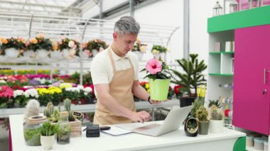 Beyaz bir çiçekçi bir saksı bitkisinin fotoğrafını çekiyor. Çiçeklerle dolu parlak bir dükkanda çiçek sergiliyor..