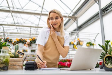 Önlüklü beyaz bir kadın çiçekçi dükkanındaki bir masada telefon kullanıyor. Konuşurken bir şeyler yazıyor..