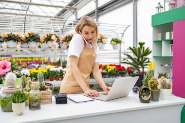 Beyaz bir kadın çiçekçide telefonla konuşurken dizüstü bilgisayarda çalışıyor. Önlük takıyor ve etrafını çiçekler ve bitkiler sarmış..