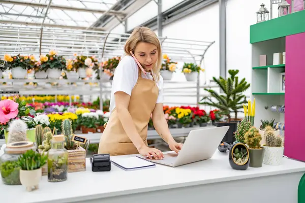 Beyaz bir kadın çiçekçide telefonla konuşurken dizüstü bilgisayarda çalışıyor. Önlük takıyor ve etrafını çiçekler ve bitkiler sarmış..