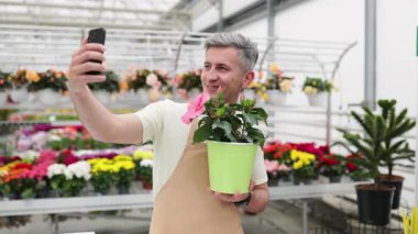 Beyaz bir çiçekçi, yeşil bir saksıda canlı pembe bir amber bitkisiyle bir çiçekçinin içinde selfie çekerken gülümsüyor..