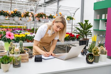 Beyaz bir kadın bir çiçekçinin satış masasında dizüstü bilgisayar kullanarak ve deftere yazı yazarak çalışıyor..
