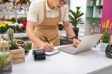 Dükkanında çalışan beyaz bir çiçekçi, bitkiler ve çiçeklerle çevrili bir dizüstü bilgisayar kullanarak not alıyor..