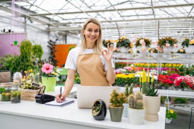 Gülümseyen bir çiçekçi dükkanında çiçek ve çiçeklerle çevrili bir şekilde satış tezgahında dururken 