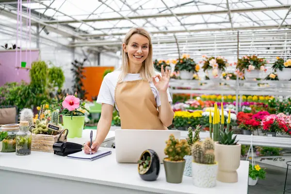 Gülümseyen bir çiçekçi dükkanında çiçek ve çiçeklerle çevrili bir şekilde satış tezgahında dururken 