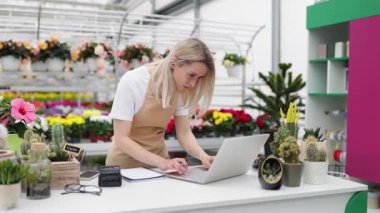 Beyaz bir kadın bir çiçekçinin satış masasında dizüstü bilgisayar kullanarak ve deftere yazı yazarak çalışıyor..