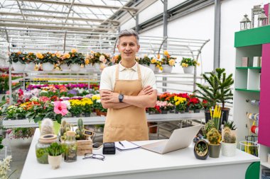 Çiçekçi dükkanında beyaz bir adam masanın arkasında çeşitli bitkilerle kameraya gülümsüyor..