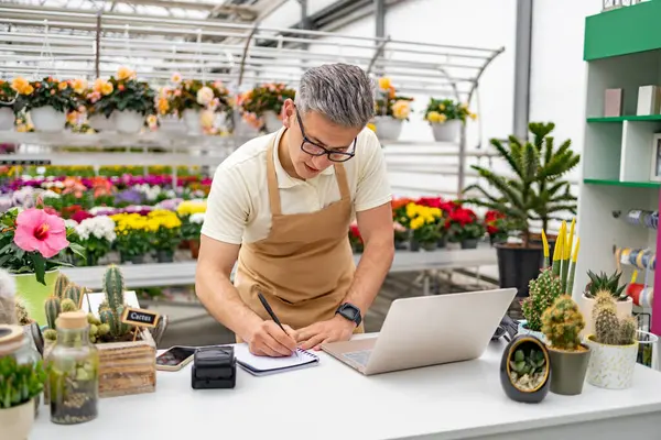 Çiçekçide çalışan beyaz bir adam. Etrafı bitki ve çiçeklerle çevrili..