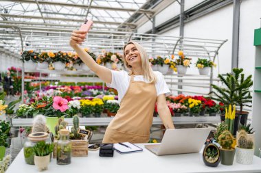 Gülümseyen beyaz bir kadın, canlı bitkilerle çevrili parlak, iyi stoklanmış çiçekçisinde selfie çekiyor..