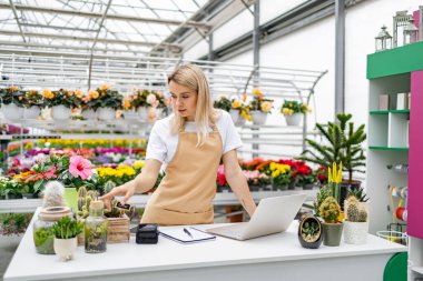 Bir çiçekçide çalışan beyaz bir kadın var. Etrafı çiçek ve bitkilerle çevrili bir dizüstü bilgisayarla bir masada duruyor, muhtemelen satışları yönetiyor..