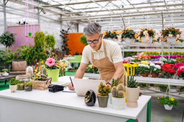 Önlük giyen beyaz bir adam çiçekçi dükkanındaki bir masada çalışıyor. Etrafı çeşitli bitki ve çiçeklerle çevrili ve dizüstü bilgisayar kullanıyor..