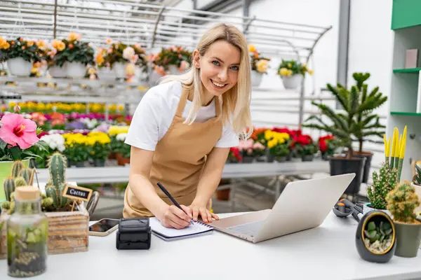 Gülümseyen bir çiçekçi bir masada dizüstü bilgisayarla çalışır ve çeşitli çiçek ve bitkilerle çevrili canlı bir dükkanda notlar alır..