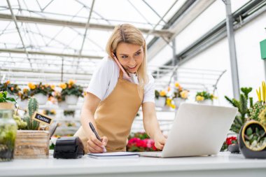 Beyaz bir kadın, çiçekçide çalışıyor, telefonla konuşuyor, notlar alıyor ve parlak bir günde dizüstü bilgisayar kullanıyor..