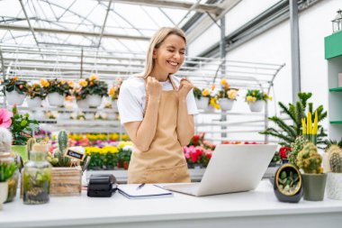 Çiçekçide neşeli bir beyaz kadın tezgahta bir satışı kutluyor. Yanında dizüstü bilgisayar, çiçekler ve bitkiler var..