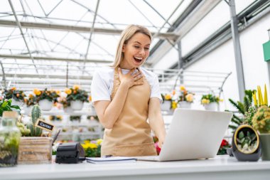 Bir çiçekçi dükkanında çiçeklerle çevrili bir dizüstü bilgisayara bakarken heyecanlanır..