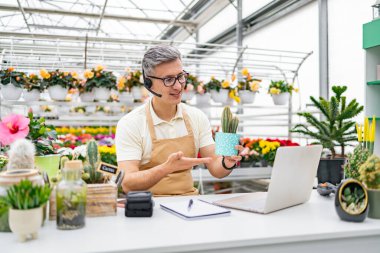 Kafkasyalı bir çiçekçi, satılık bir kaktüsü sergilerken bir video görüşmesi yapıyor ve bir dizüstü bilgisayarla online satışları vurguluyor..