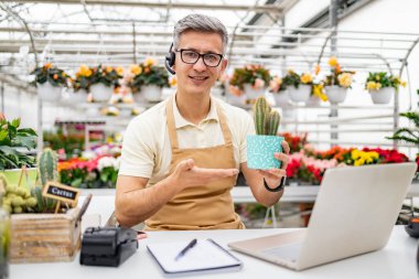 Çiçekçi dükkanındaki beyaz bir adam, kameraya gülümseyen ve el kol hareketi yapan bir video görüşmesinde elinde bir kaktüs tutuyor..