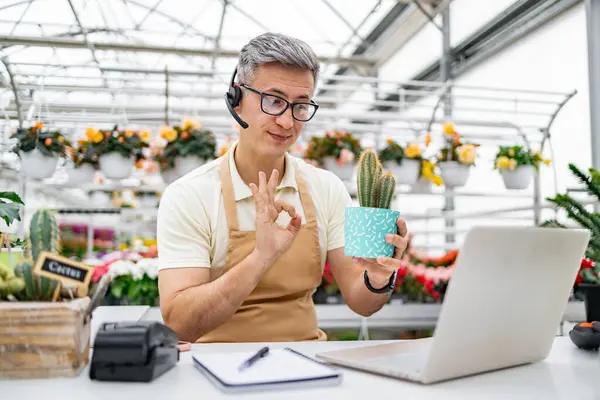 Çiçekçi dükkanındaki beyaz bir adam video görüşmesi sırasında kaktüs gösteriyor. Santrali tutuyor ve sinyal veriyor..