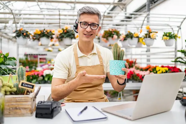 Çiçekçi dükkanındaki beyaz bir adam, kameraya gülümseyen ve el kol hareketi yapan bir video görüşmesinde elinde bir kaktüs tutuyor..
