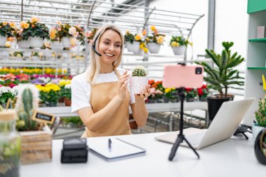 Bir kadın, çiçekçide canlı satış sırasında telefonuyla çekim yaparken kaktüs gösteriyor..