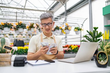 Çiçekçi dükkanındaki beyaz bir adam kazandıklarını sayıyor. Etrafı renkli çiçekler ve satılık bitkilerle çevrili. Tezgahta..
