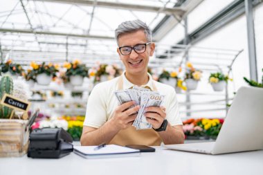 Kafkasyalı bir çiçekçi alışveriş masasında yığınla para tutuyor, satışlardan memnun bir şekilde gülümsüyor. Dükkanda çiçekler var..