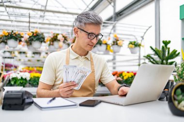 Elinde nakit para olan beyaz bir çiçekçi çiçekçide dizüstü bilgisayar kullanıyor. Satış ve finans işlerini yürütüyor..