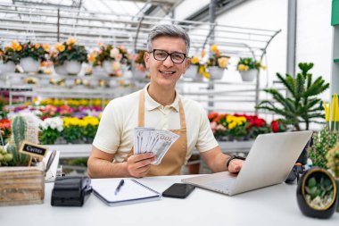 Gülümseyen beyaz bir adam elinde para tutuyor, güzel bitkilerle çevrili bir çiçekçide dizüstü bilgisayarla çalışıyor..