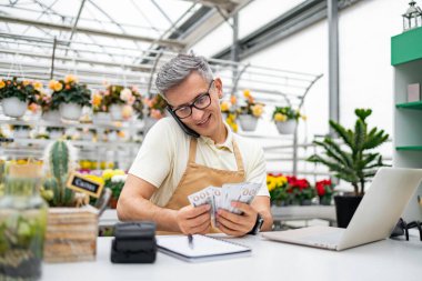 Beyaz bir çiçekçi, telefonda çiçekçinin içindeki satış masasında para sayıyor. Başarılı bir satış işareti..